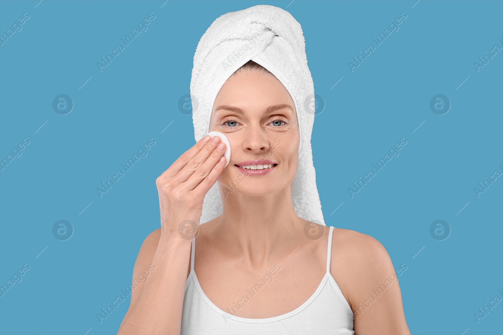 Photo of Beautiful woman in terry towel removing makeup with cotton pad on light blue background