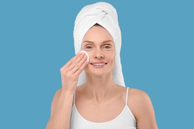 Beautiful woman in terry towel removing makeup with cotton pad on light blue background