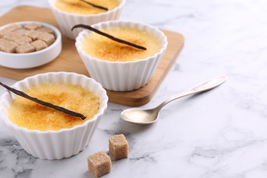 Photo of Delicious creme brulee in bowls, vanilla pods, sugar cubes and spoon on white marble table, closeup. Space for text
