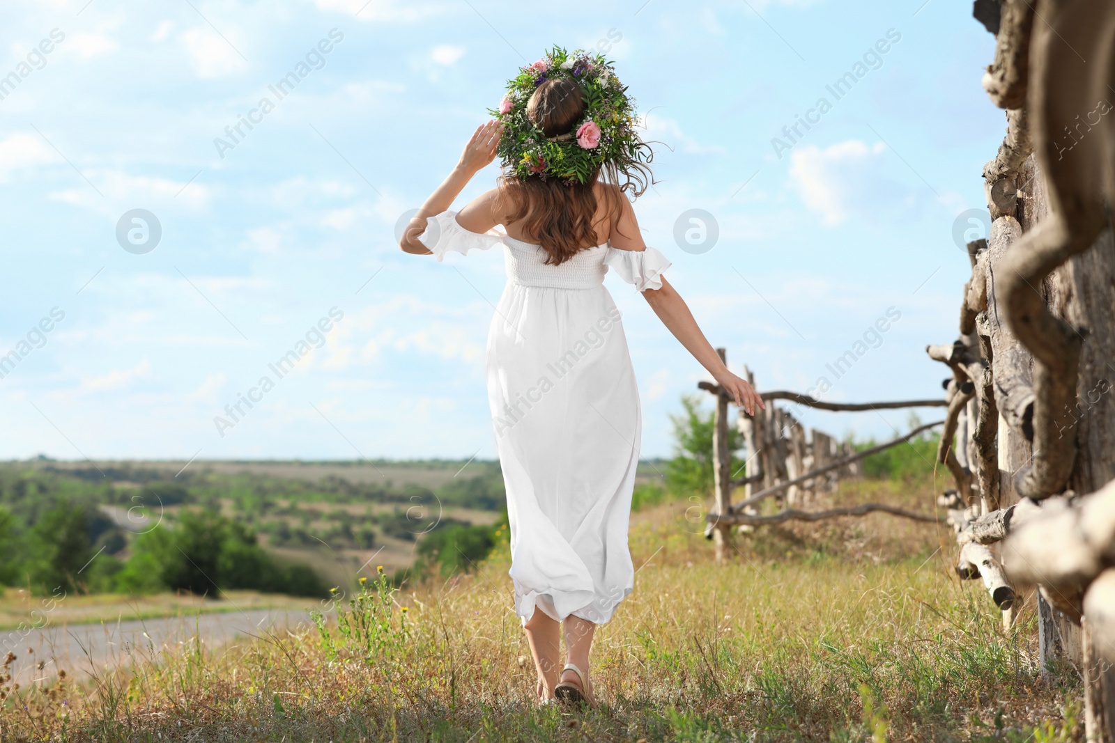 Photo of Young woman wearing wreath made of beautiful flowers outdoors on sunny day, back view