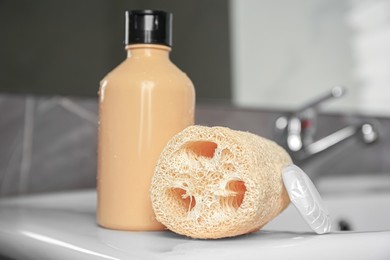 Photo of Natural loofah sponge and shower gel bottle on washbasin in bathroom, closeup