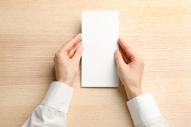 Young woman holding blank brochure at light wooden table, top view. Mock up for design