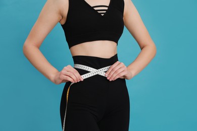 Woman in sportswear measuring waist with tape on light blue background, closeup