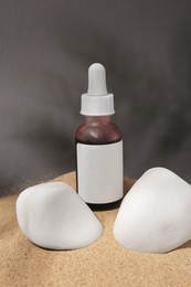 Photo of Bottle of serum and stones on sand against grey background