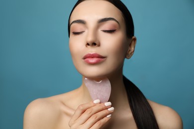 Photo of Beautiful young woman doing facial massage with gua sha tool on blue background, closeup