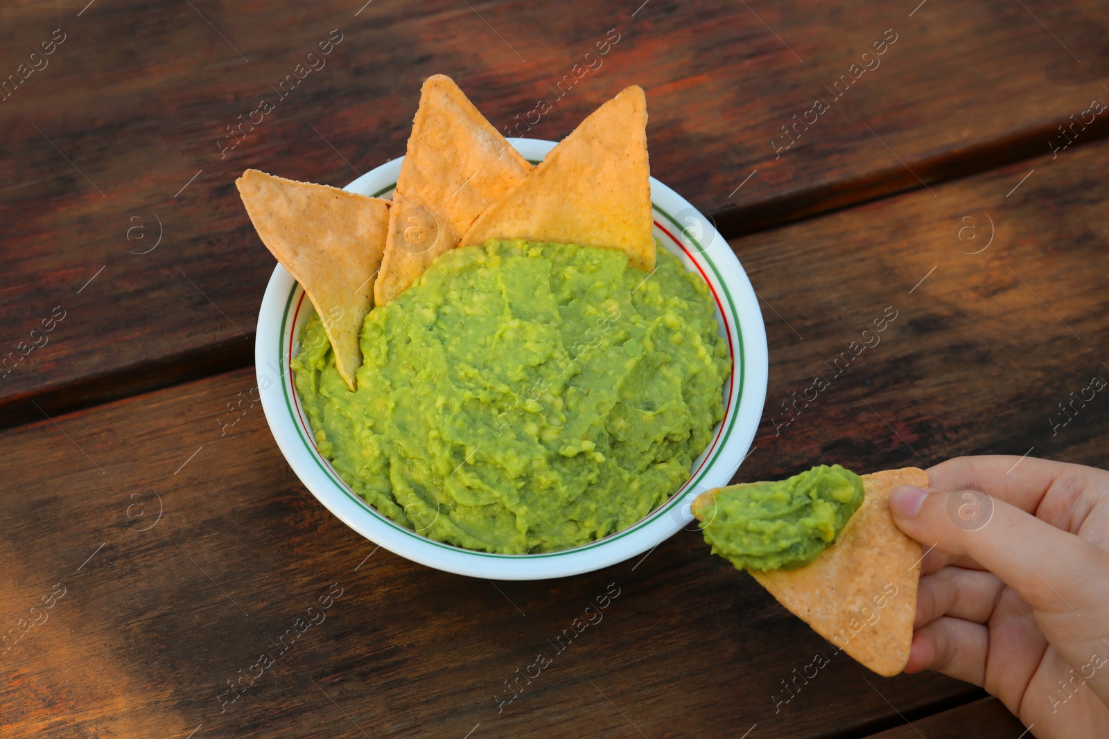 Photo of Woman holding nacho chip with delicious guacamole made of avocados at wooden table, closeup