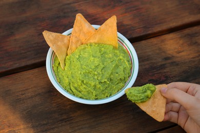 Photo of Woman holding nacho chip with delicious guacamole made of avocados at wooden table, closeup