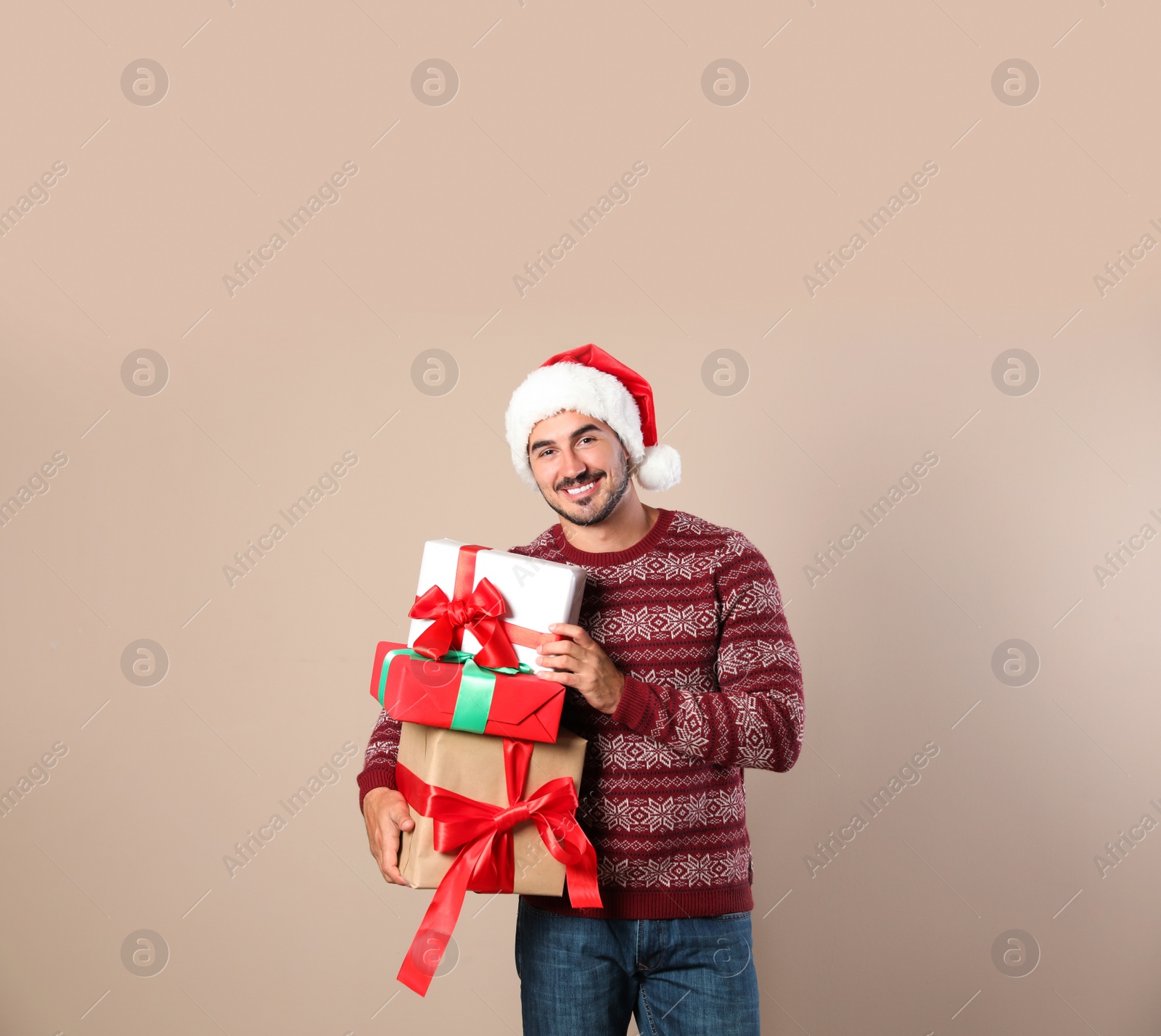 Photo of Happy man in Christmas sweater and Santa hat holding gift boxes on beige background