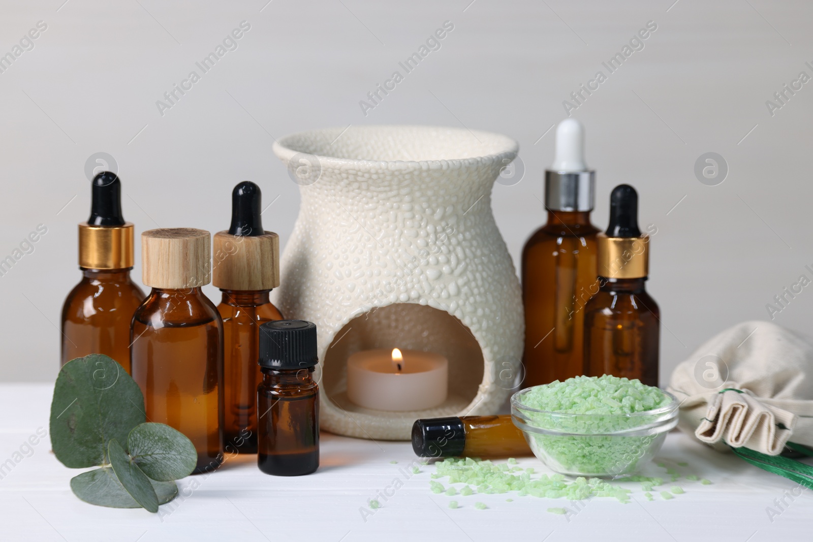 Photo of Different aromatherapy products, burning candle and eucalyptus leaves on white wooden table against light background