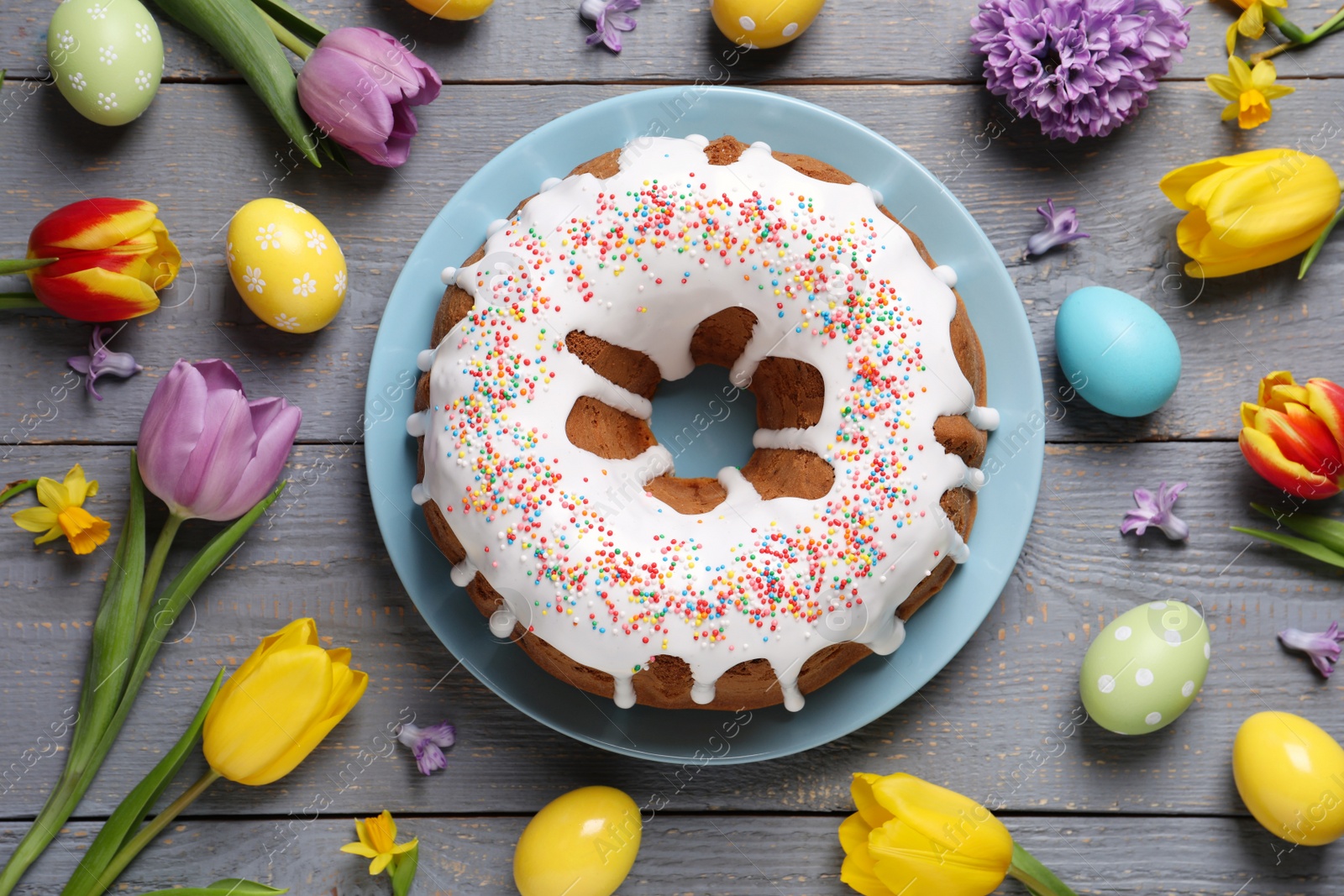 Photo of Glazed Easter cake with sprinkles, painted eggs and flowers on grey wooden table, flat lay