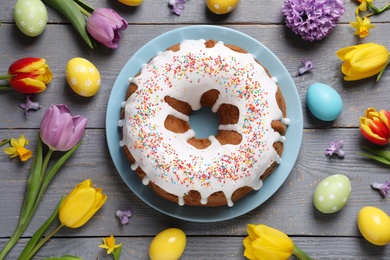 Glazed Easter cake with sprinkles, painted eggs and flowers on grey wooden table, flat lay