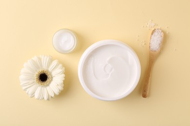 Photo of Moisturizing cream in open jars, sea salt and gerbera flower on beige background, flat lay. Body care product