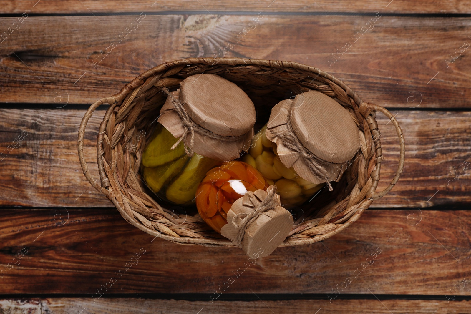 Photo of Wicker basket with many jars of different preserved products on wooden table, top view