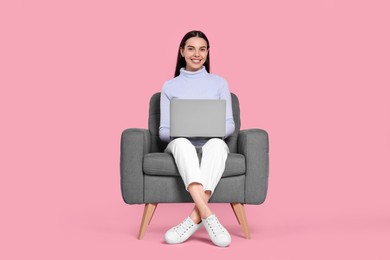 Happy woman with laptop sitting in armchair on pink background