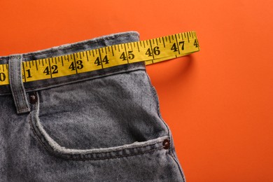 Jeans and measuring tape on orange background, top view with space for text. Weight loss concept
