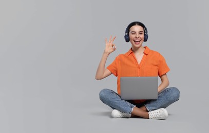 Photo of Happy woman with laptop wearing headphones and showing OK gesture on light gray background