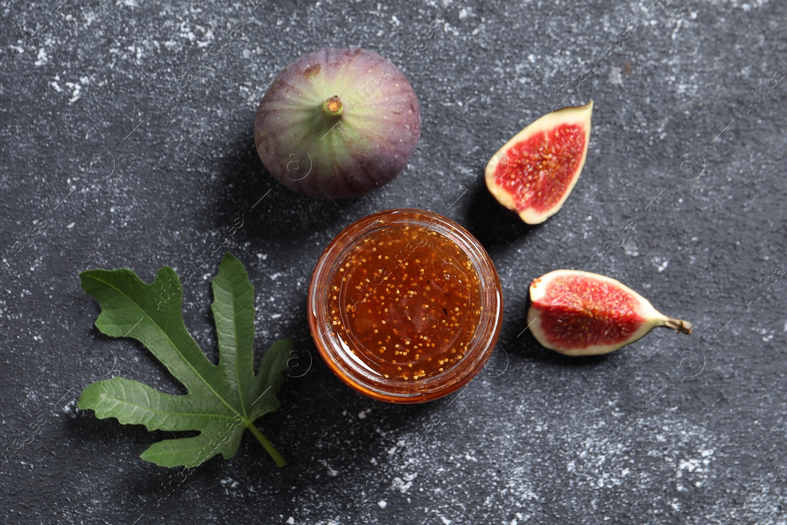 Photo of Jar with tasty sweet jam, fresh figs and green leaf on black textured table, flat lay
