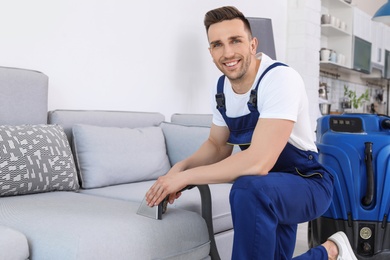 Male worker removing dirt from sofa with professional vacuum cleaner indoors