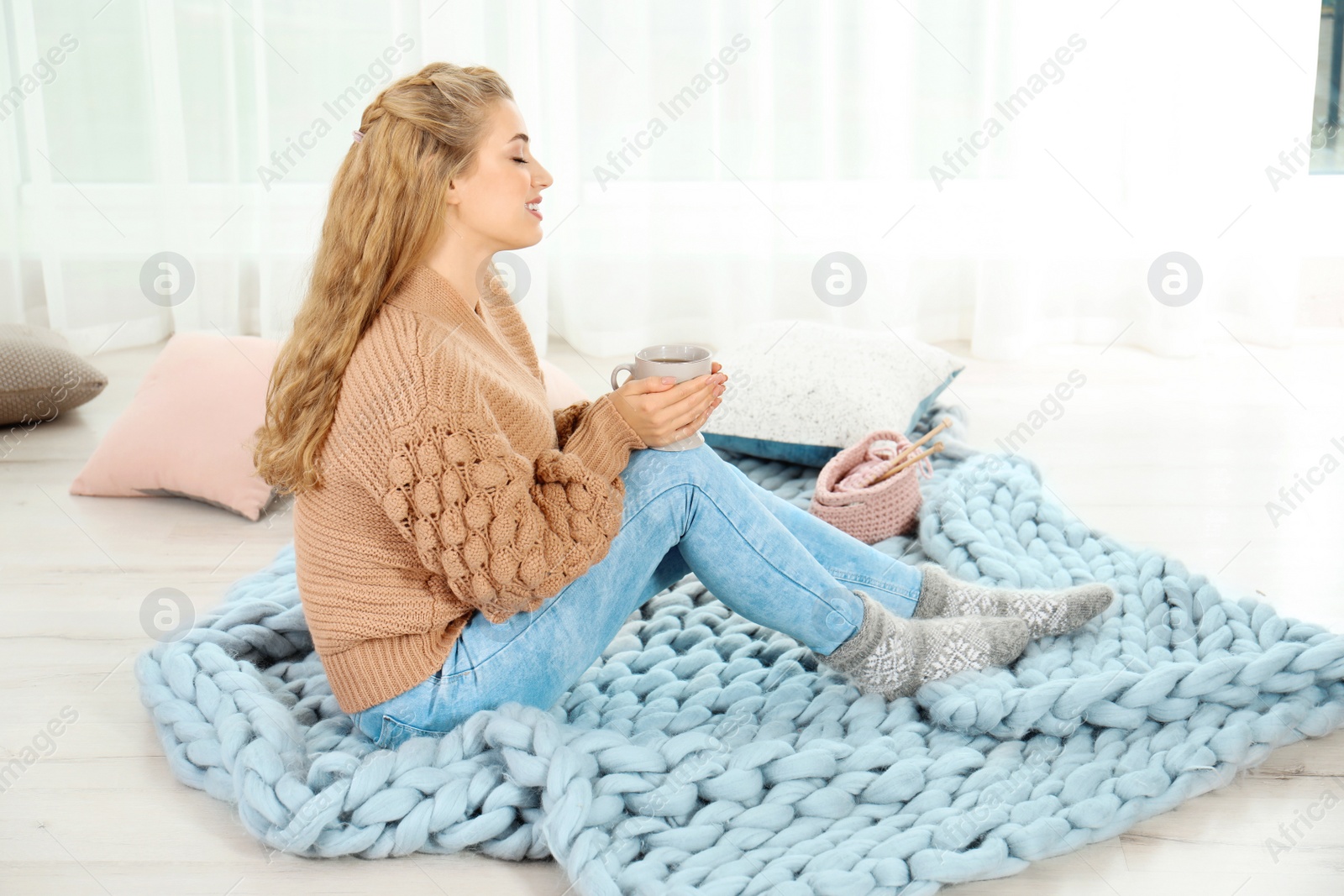 Photo of Attractive young woman in cozy warm sweater with cup of hot drink sitting on floor at home. Space for text