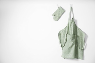 Photo of Clean apron with pattern, kitchen tools and oven glove on light wall. Space for text