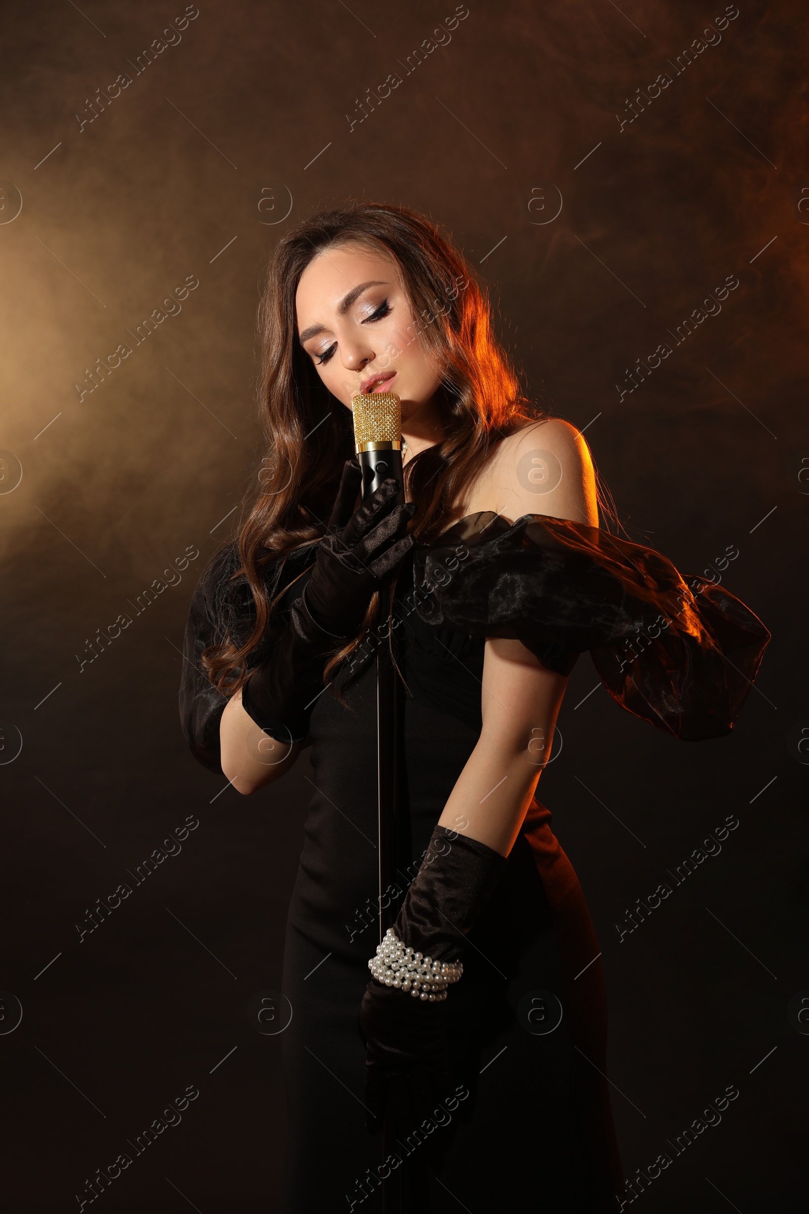 Photo of Beautiful young woman with microphone singing on dark background with smoke