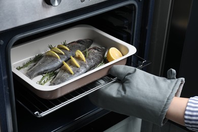 Photo of Woman putting baking tray with sea bass fish, lemon and rosemary into oven, closeup