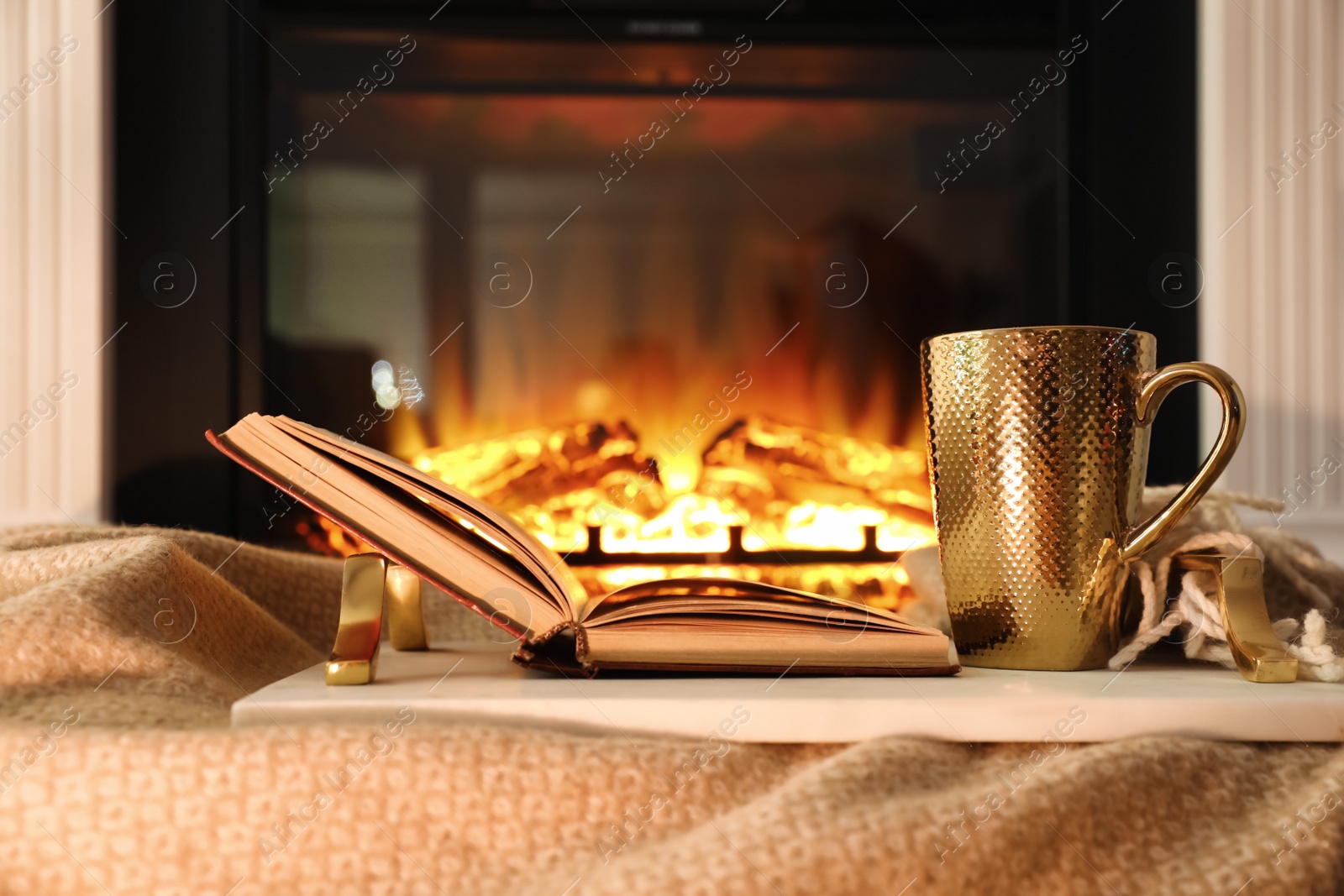 Photo of Cup of hot drink and book on blanket near fireplace indoors. Magic atmosphere