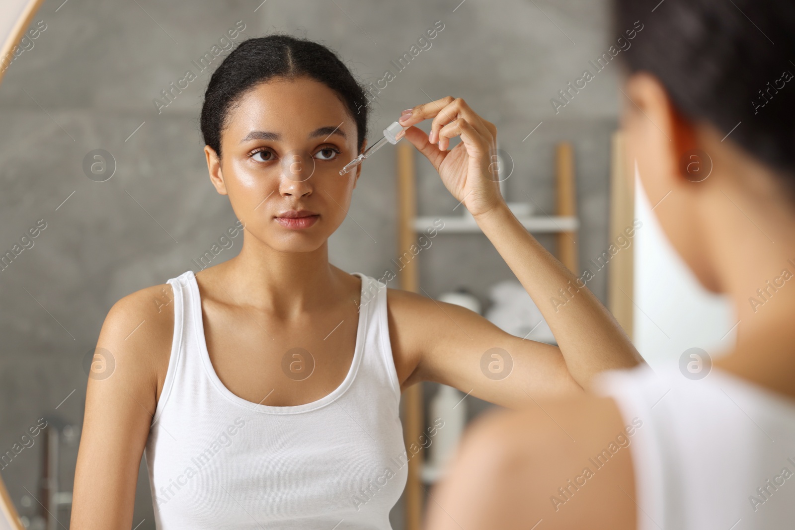 Photo of Beautiful woman applying serum onto her face near mirror in bathroom