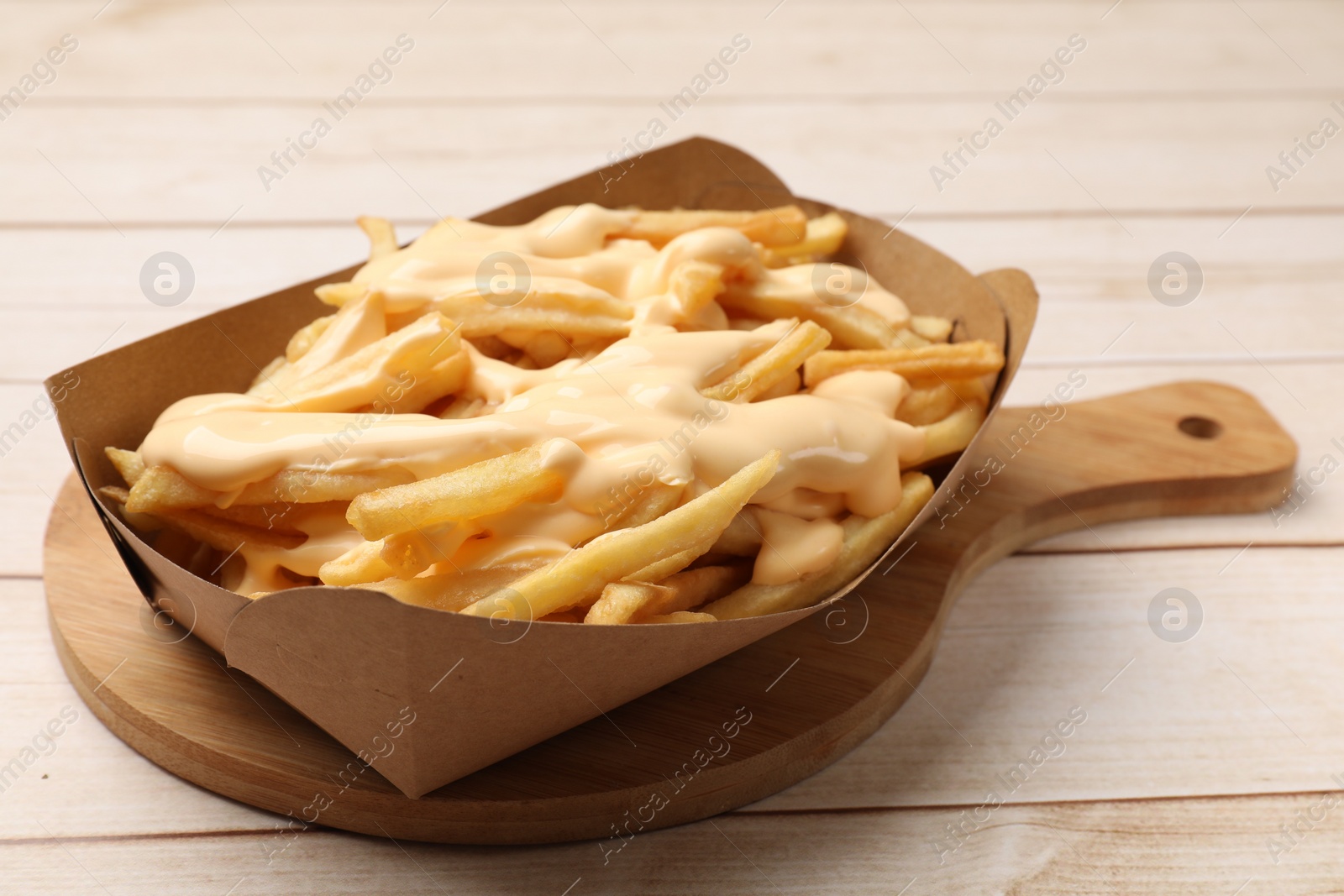 Photo of Tasty potato fries and cheese sauce in paper container on light wooden table, closeup