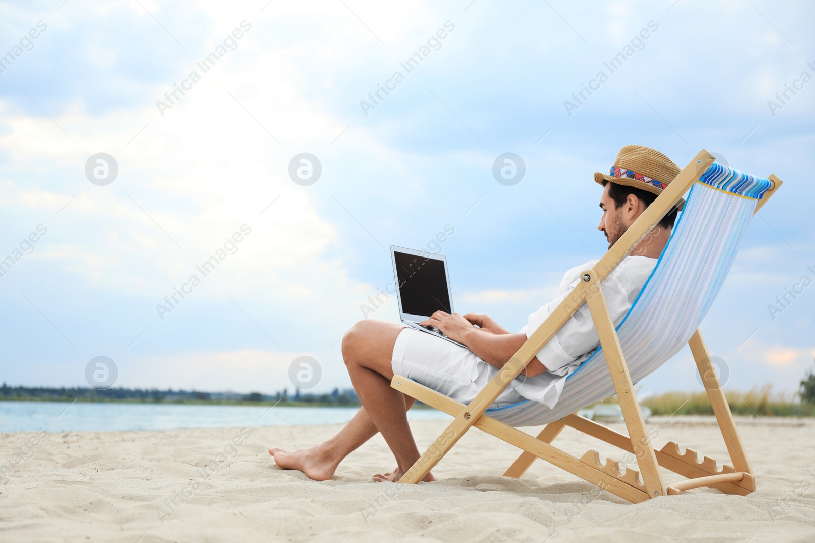 Photo of Young man with laptop sitting in deck chair on sea beach. Space for text