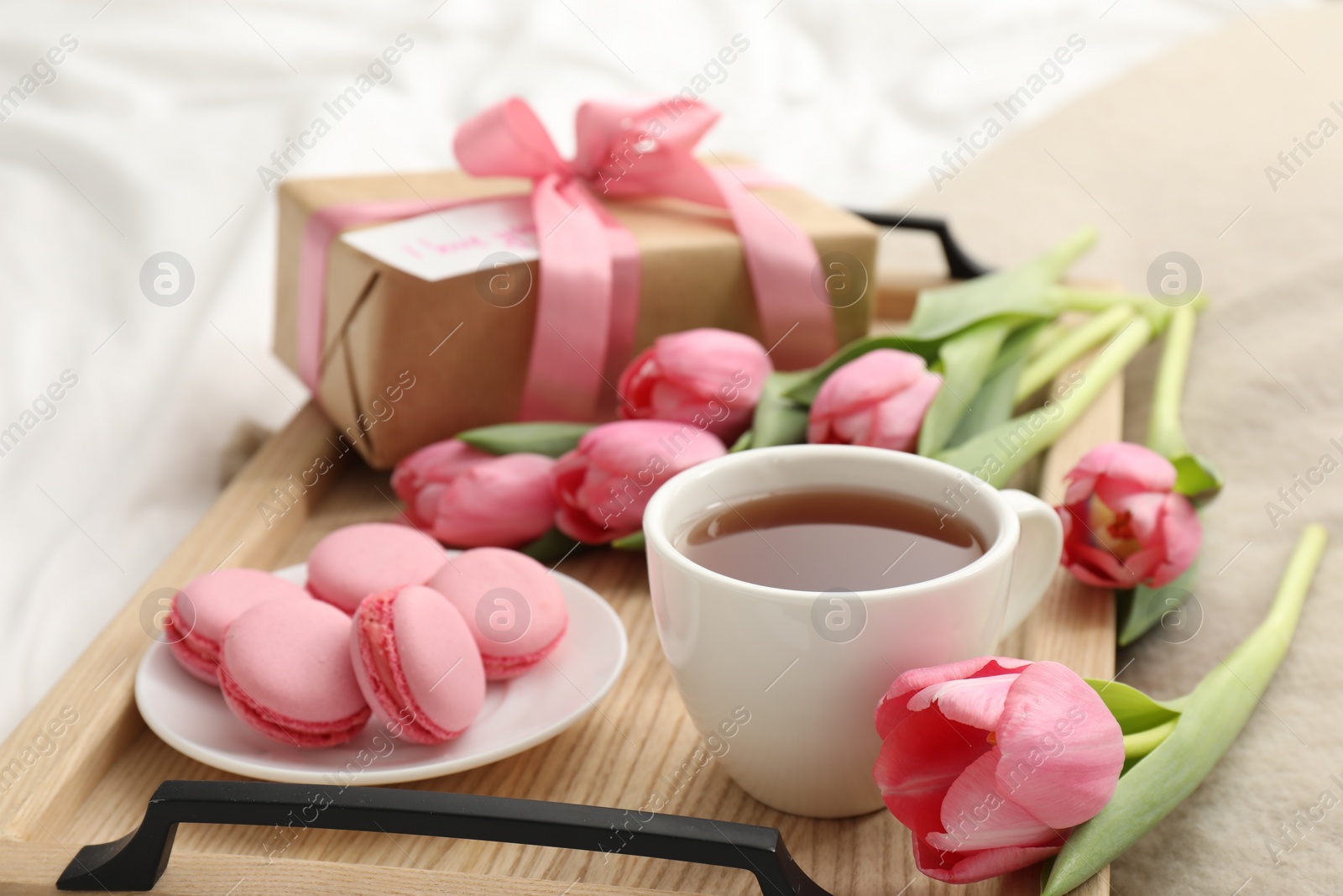 Photo of Tasty breakfast served in bed. Delicious macarons, tea, flowers and gift box on tray, closeup