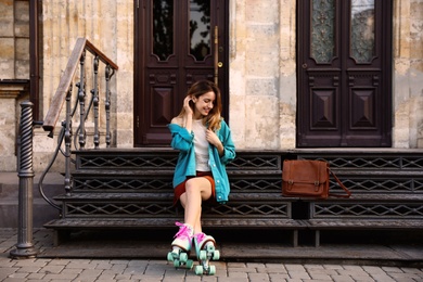 Photo of Happy stylish young woman with vintage roller skates and bag sitting on stairs outdoors