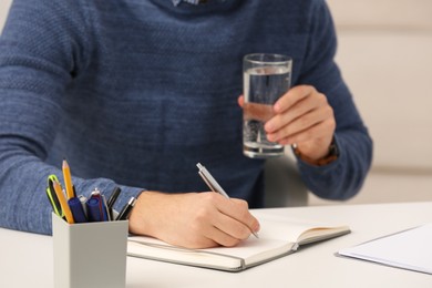 Man with glass of water writing in notebook at table indoors, closeup
