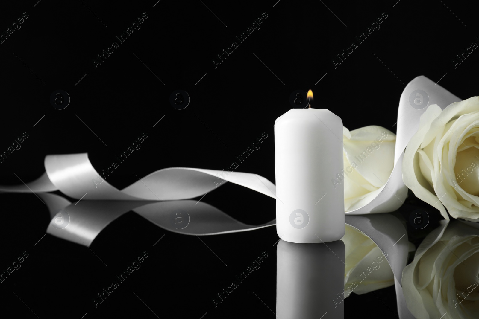 Photo of Burning candle, white roses and ribbon on black mirror surface in darkness, space for text. Funeral symbols