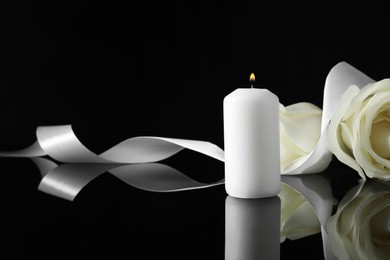 Photo of Burning candle, white roses and ribbon on black mirror surface in darkness, space for text. Funeral symbols