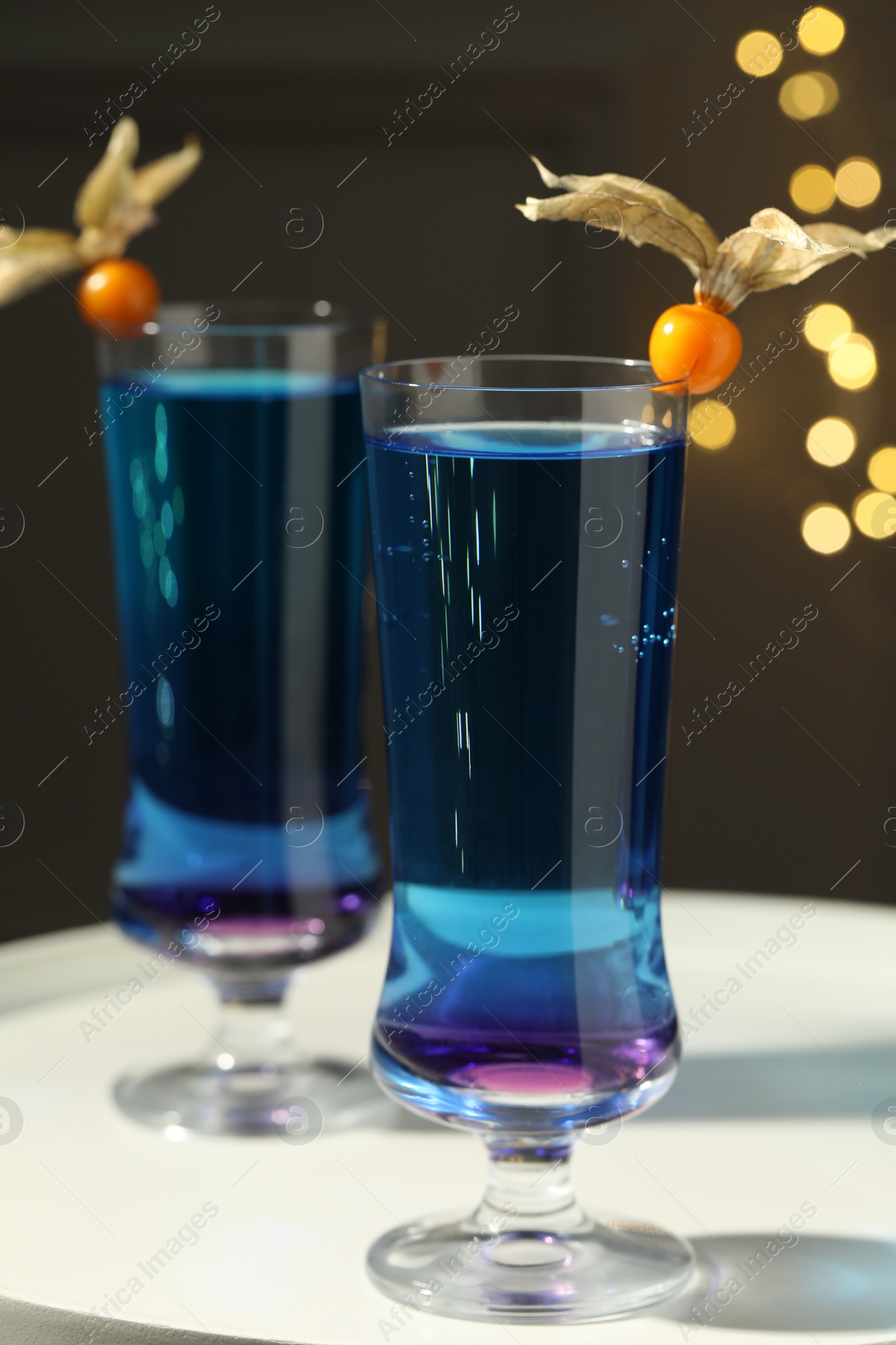 Photo of Refreshing cocktails decorated with physalis fruits on white table against blurred lights