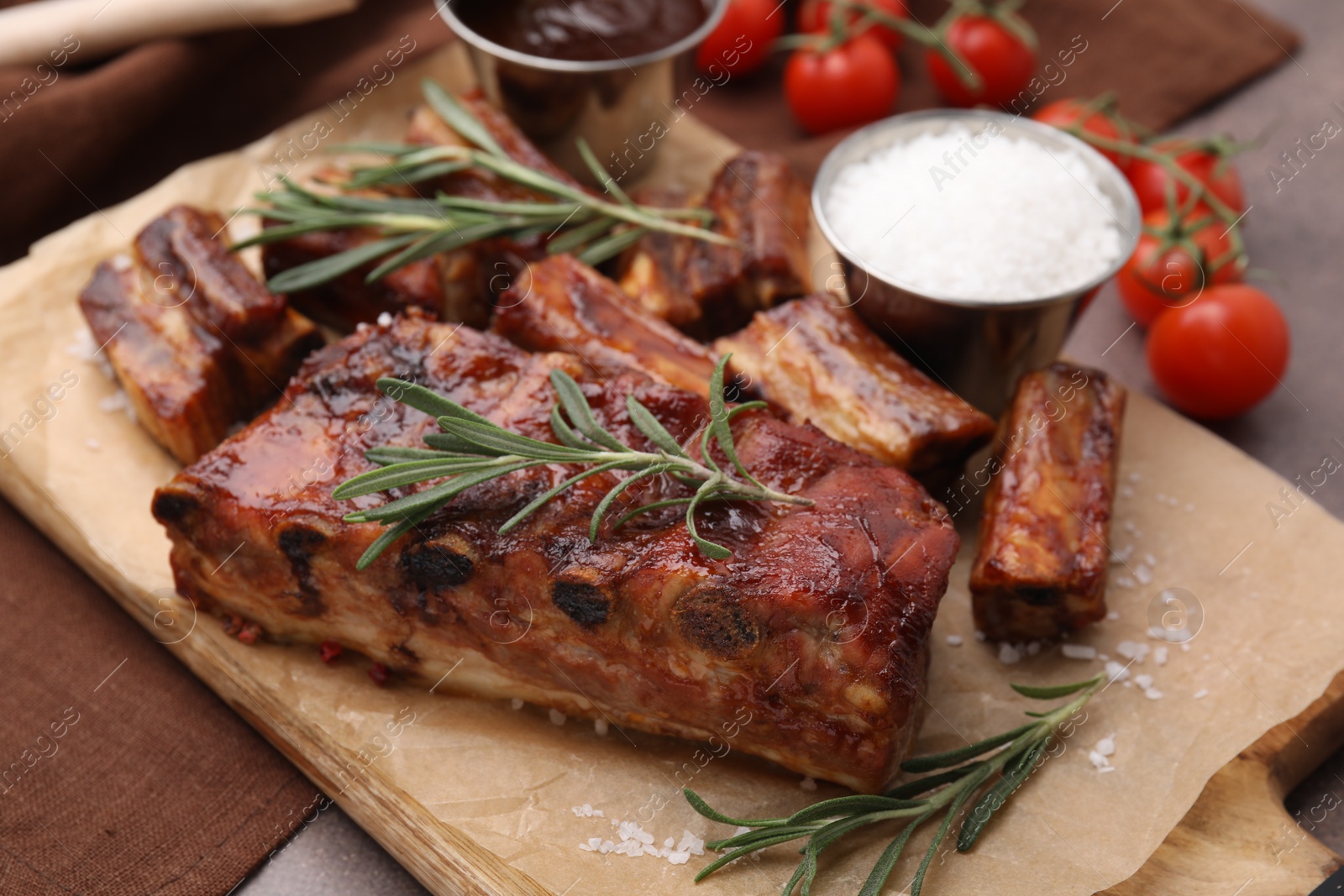 Photo of Tasty roasted pork ribs served with sauce, rosemary and tomatoes on brown table, closeup