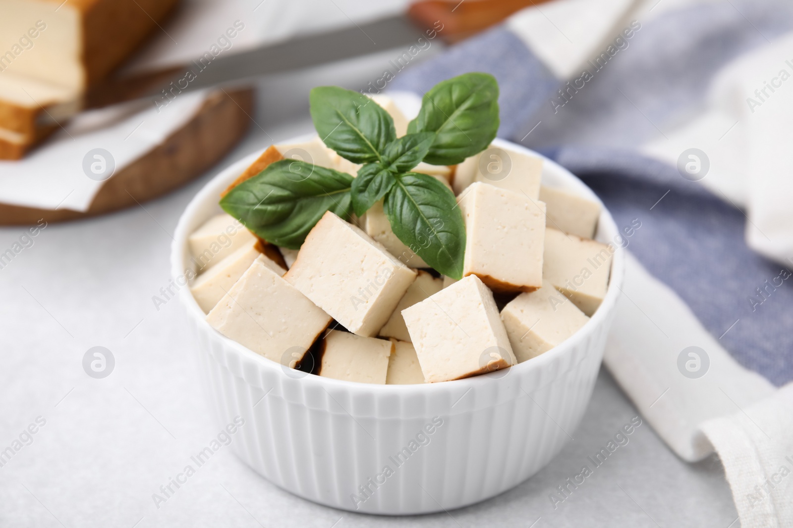 Photo of Bowl of smoked tofu cubes with basil on white table, closeup