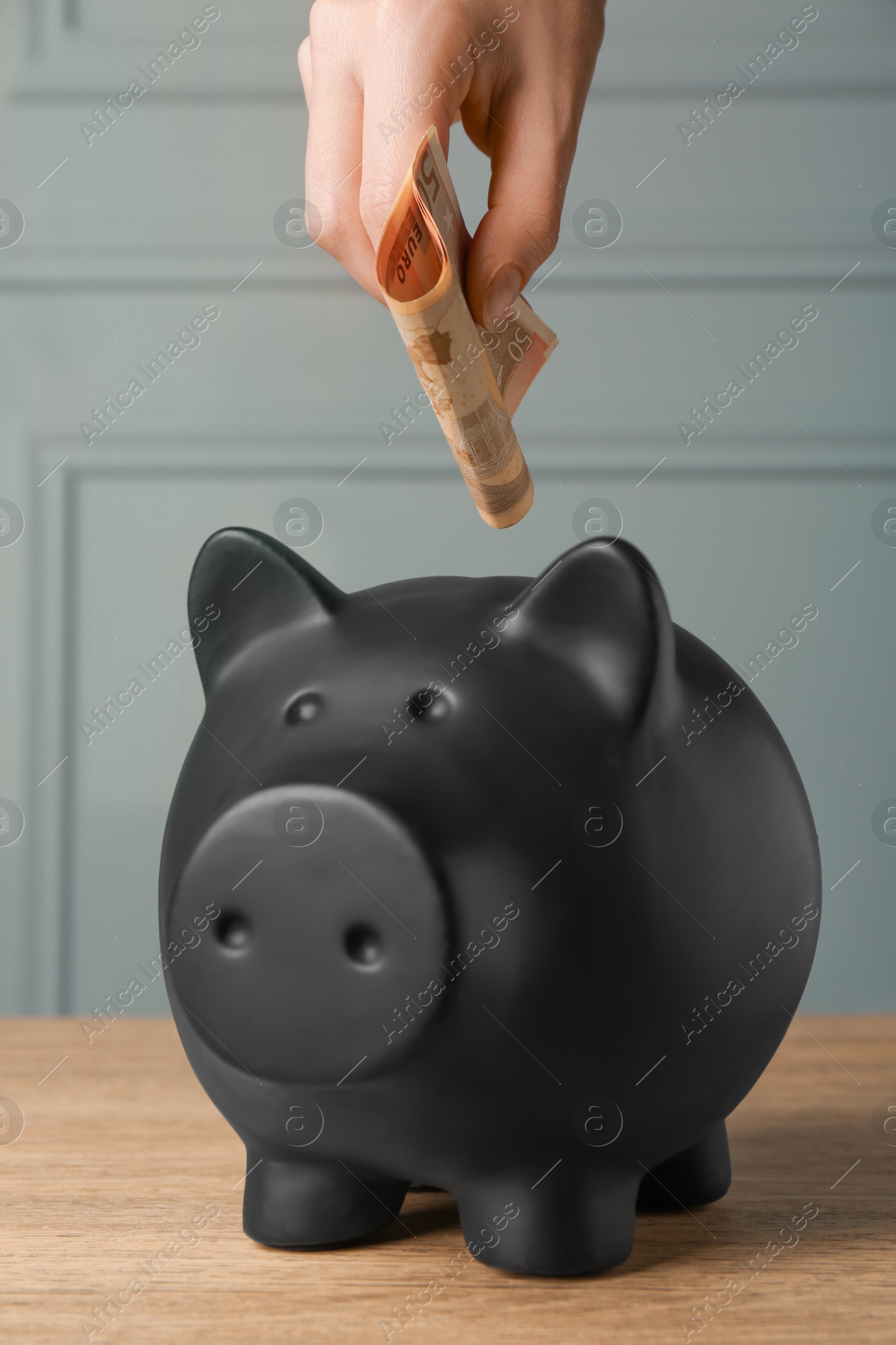 Photo of Woman putting banknotes into piggy bank at wooden table, closeup