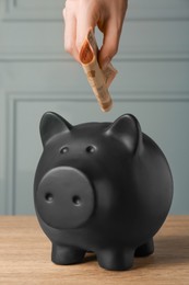 Photo of Woman putting banknotes into piggy bank at wooden table, closeup