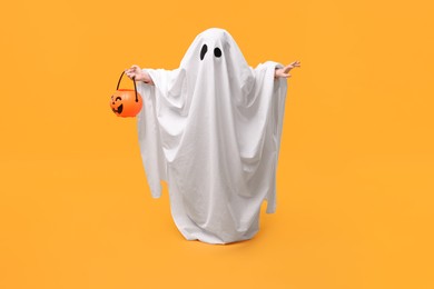 Child in white ghost costume holding pumpkin bucket on orange background. Halloween celebration