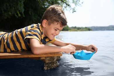 Photo of Cute little boy playing with paper boat on wooden pier near river