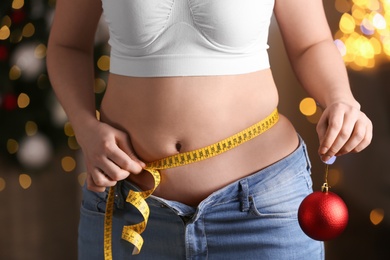 Photo of Woman with Christmas ball and measuring tape indoors, closeup. Overweight problem after New Year party