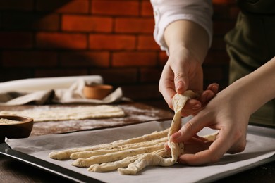 Photo of Woman putting homemade breadsticks on baking sheet at wooden table indoors, closeup. Cooking traditional grissini