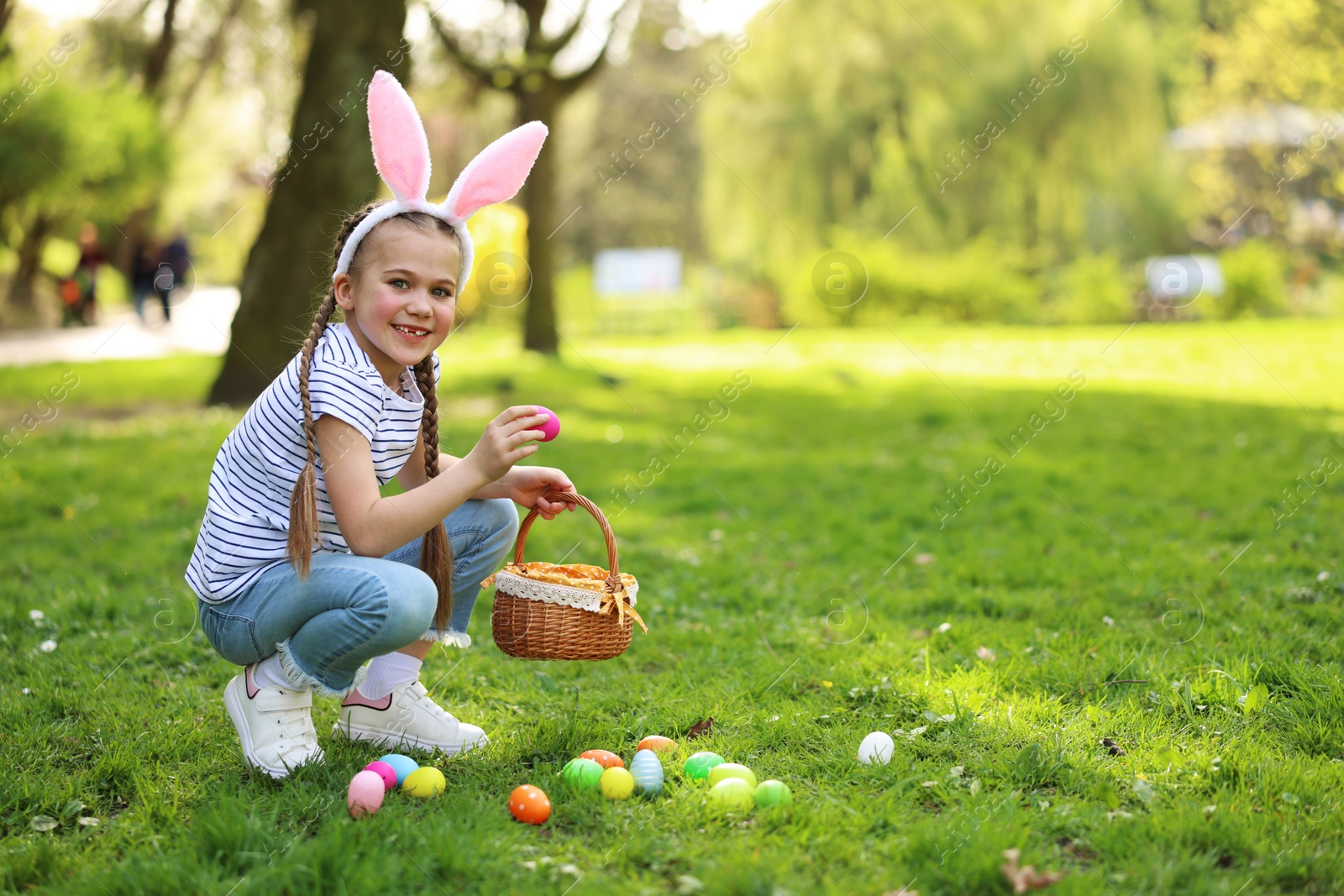 Photo of Easter celebration. Cute little girl in bunny ears hunting eggs outdoors, space for text