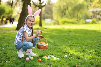 Easter celebration. Cute little girl in bunny ears hunting eggs outdoors, space for text