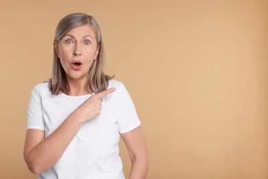 Surprised senior woman pointing at something on beige background, space for text