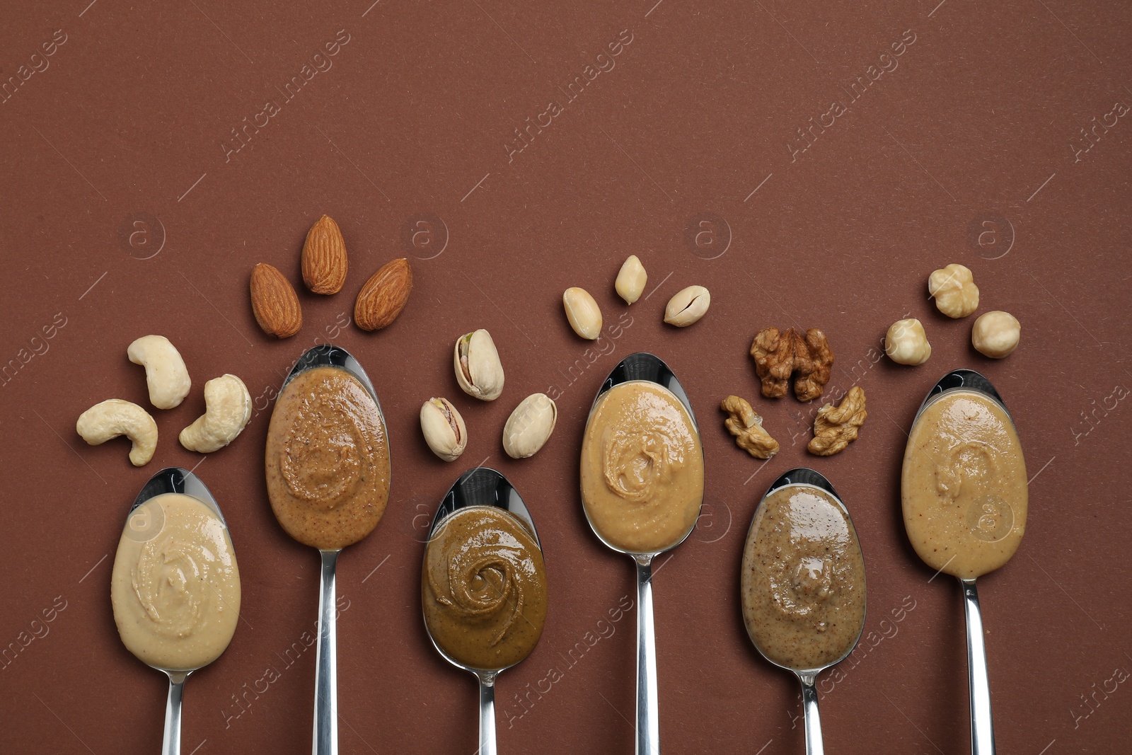 Photo of Tasty nut butters in spoons and raw nuts on brown table, flat lay. Space for text