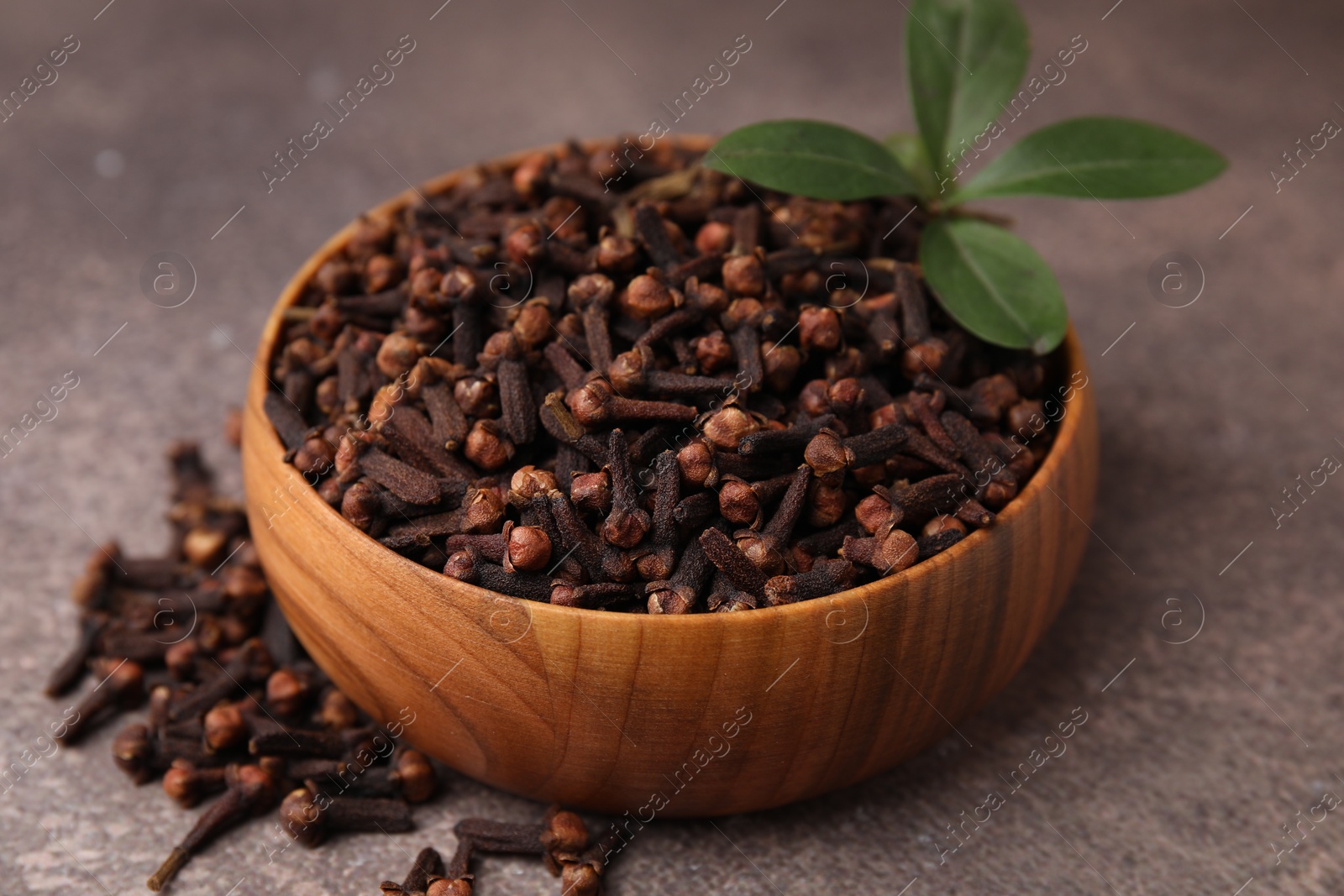 Photo of Aromatic cloves and green leaves in bowl on brown table, closeup