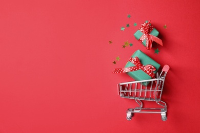 Photo of Small shopping cart with gift boxes on red background, flat lay. Space for text